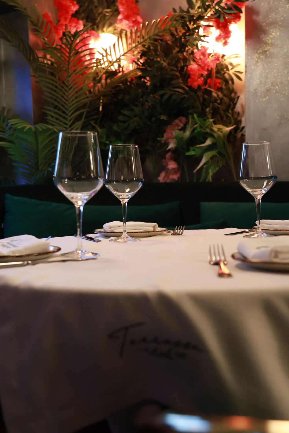 A table laid with embroidered linen beside the green velvet banquette and flower wall